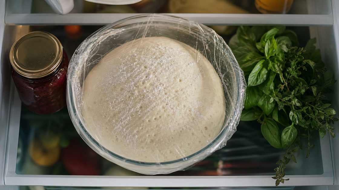 Gerezen pizzadeeg in een kom afgedekt met folie op een plank in de koelkast