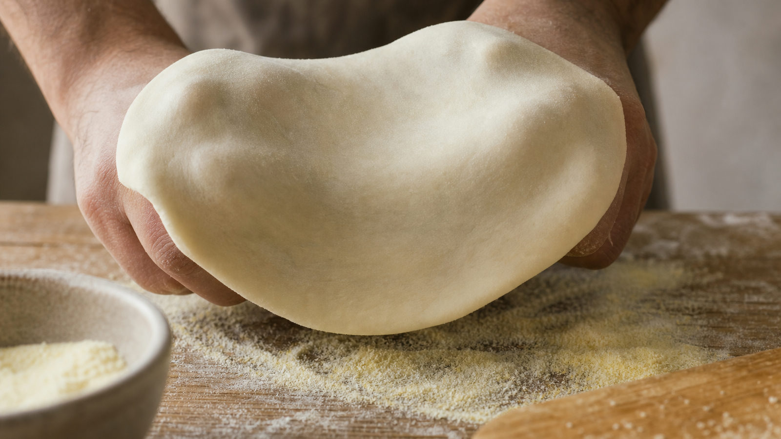 Twee handen rekken een ronde pizzadeegbol uit boven een houten werkblad met semola