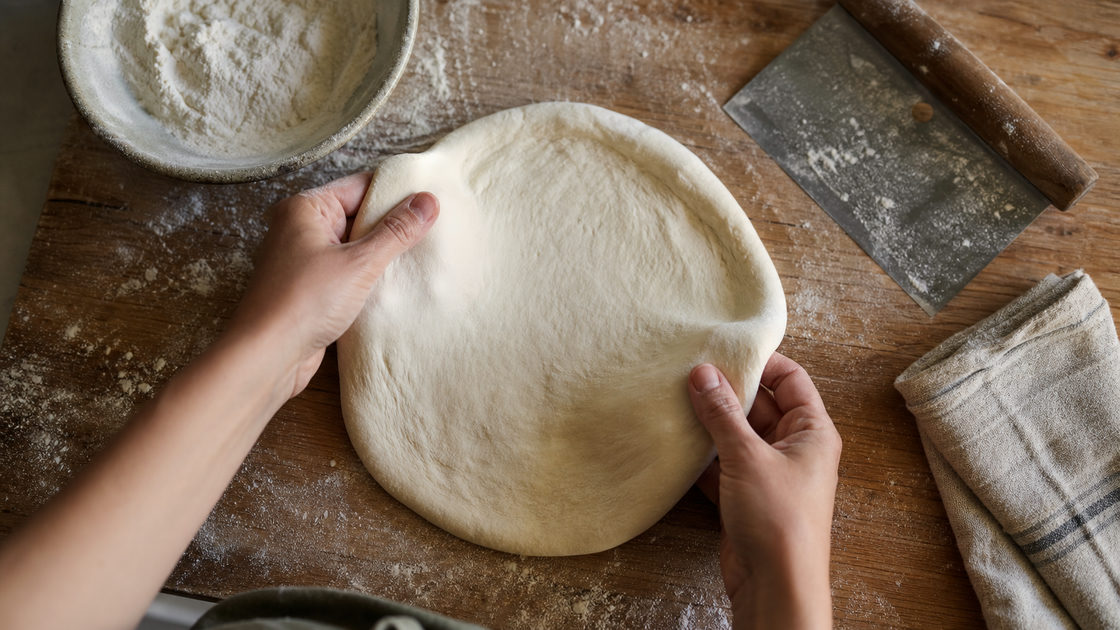 Handen die een ronde pizzadeegbol uitrekken op een bebloemd houten werkblad met een schaaltje bloem ernaast