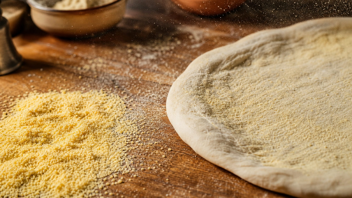 Grof geel semolina meel uitgestrooid op een houten werkblad naast een uitgerekte bol pizzadeeg