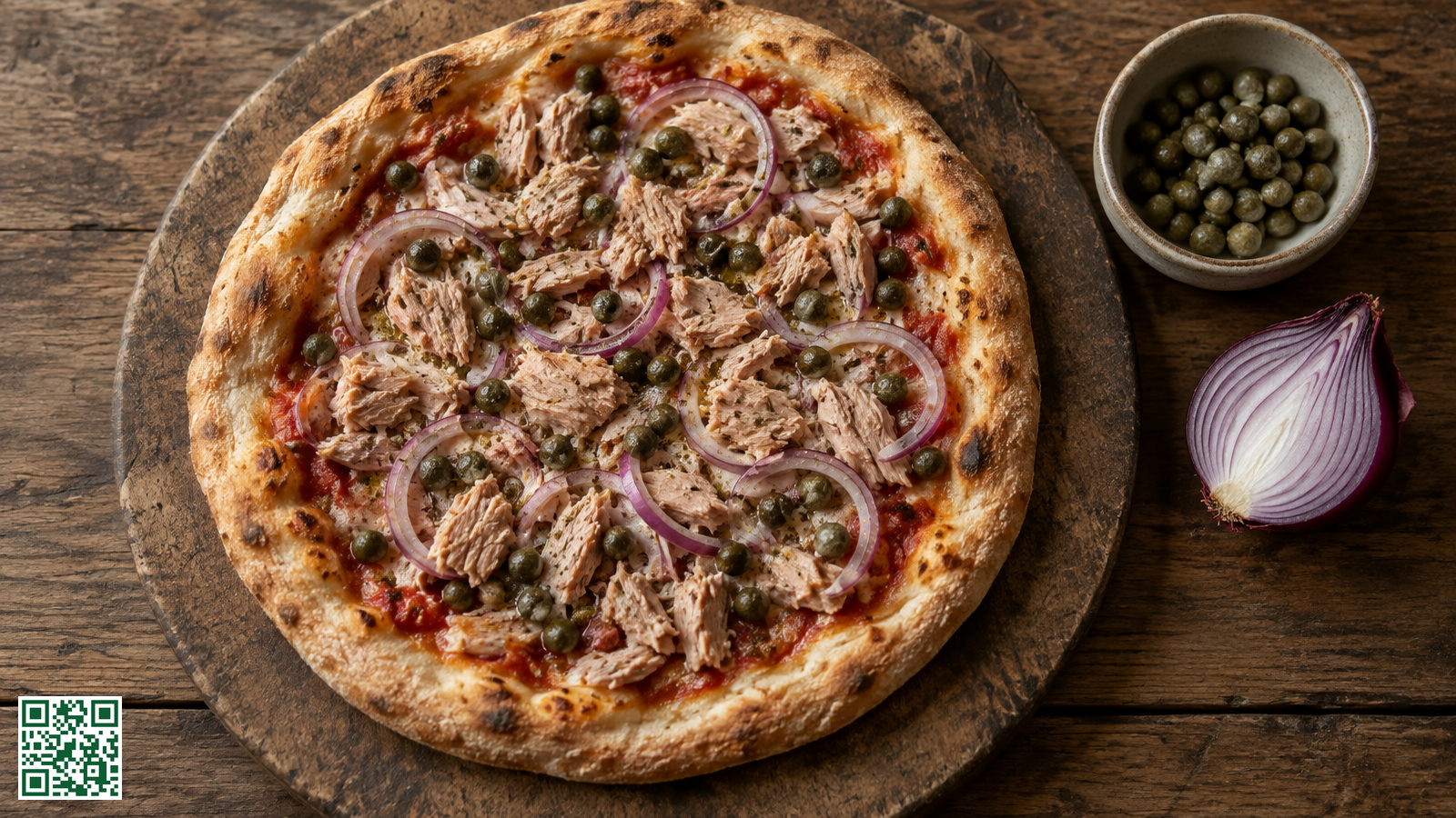 Pizza met stukjes tonijn, ringen rode ui en kappertjes op een houten plank